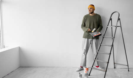 a young man of European appearance stands near the wall. in the hands of a roller and a tray for paint. Copy space.の写真素材