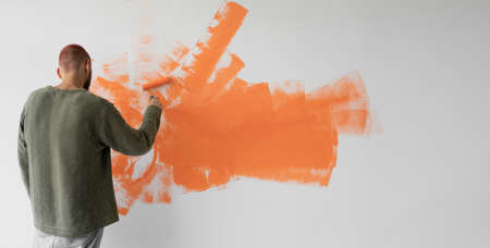 a young man with a roller in his hands paints the wall with orange paint. Copy space. Home renovation conceptの写真素材