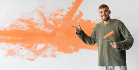a young man of European appearance stands near the wall. Holds a paint roller. points her finger at an empty space for text. Home renovation concept. Baner. Copy spaceの写真素材