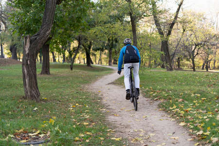 A man with a backpack rides a bicycle through the park on a bicycle road. He has a safety helmet and jacket. Place for text.の写真素材