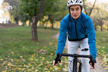 Portrait of a young sporty man who rides a bicycle through the park on a bicycle road. He has a safety helmet and jacket. Copy space.の写真素材