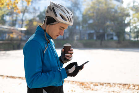 A young man in a bicycle helmet drinks hot coffee from a glass and reads orders in a smartphone. Food delivery around the city on bicycles. Copy space.の写真素材