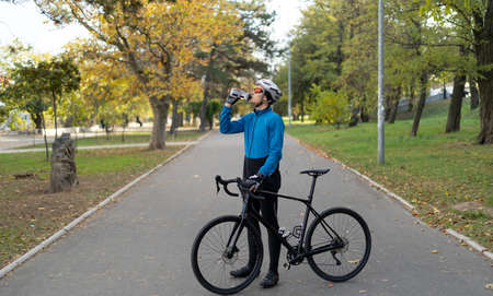 A young man in a bicycle helmet drinks water from a fitness bottle and holds a bicycle in his hands. Sports activities and food delivery around the city on bicycles. Place for text.の写真素材