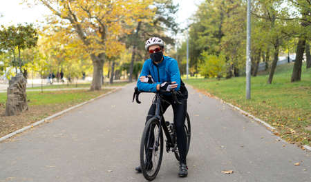 A man on a bicycle wearing safety goggles, a black mask and a bicycle helmet on a bicycle road in the park. Quarantine sports and outdoor food delivery.の写真素材