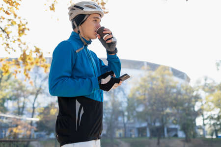 A young man in a bicycle helmet drinks hot coffee from a glass and holds a smartphone in his hands. Food delivery around the city on bicycles. Place for text.の写真素材