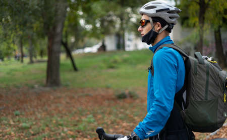 A man of Caucasian ethnicity in the park with a bicycle. Sportsman on vacation. Food delivery in quarantine and protective medical mask. Place for your text.の写真素材
