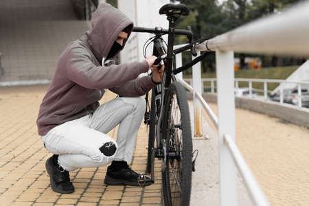A thief in a balaclava and a hood breaks the lock on a bicycle in the street during the day. Closed face and hacking. Stealing bicycles. Place for your text.の写真素材