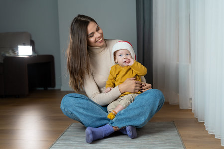 A young woman with a child in a Santa Claus hat sits on the floor in the room. New Years and...