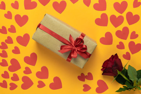 top view of a red rose lies on a yellow background. Lots of red cardboard hearts of different sizes....