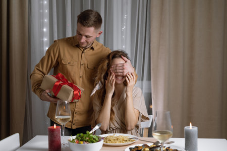 The young man makes a gift to his beloved and covers her eyes with his hand. A beautiful woman receives a surprise gift box from her boyfriend in a restaurant. Valentines Day. Womens Day.の写真素材