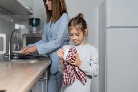 Caucasian child girl dries dishes in the kitchen, wiping with a kitchen towel. Parents help concept.の写真素材