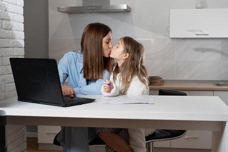 mom and daughter sit together at home at the table in the evening watching a drawing lesson on a laptop. kiss each other.の写真素材