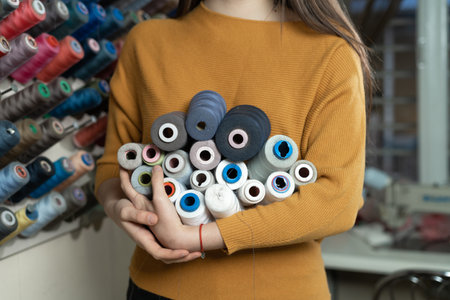 A young girl seamstress of Caucasian ethnicity in a workshop stands near a stand with threads of different colors. She holds various skeins in her hands. The choice of suture material for sewing a shirt. Copy space.の写真素材