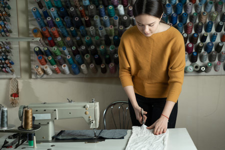 A dressmaker stands at a work table and cuts white fabric along a pattern with scissors. The...