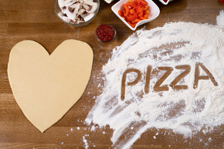 On the table in a pizzeria there is a heart-shaped pizza blank and ingredients for cooking. The flour crumbles and there is an inscription on it. Surprise for Valentines Day.の写真素材