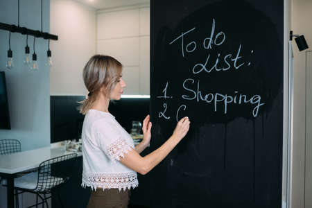 A blonde woman stands with her back and writes in chalk on a black board in the kitchen at home. To-do list in the kitchen.の写真素材