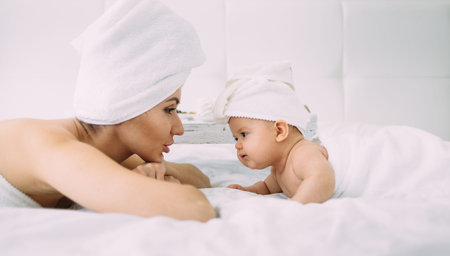 A young mother and her six-month-old daughter lie on a white bed with towels tied to their heads....