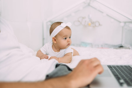 A six-month-old baby lies on a white bed and watches dad study while sitting next to a laptop.の写真素材