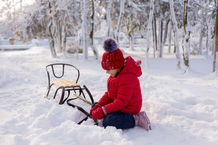 A six-year-old girl sits on her knees in the snow in a winter park with a sleigh next to her. The child is tired and sad.の写真素材