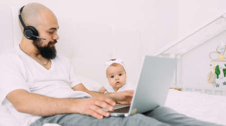 A young dad is sitting on a bed with a child together at home at a computer. A father works on a laptop.の写真素材