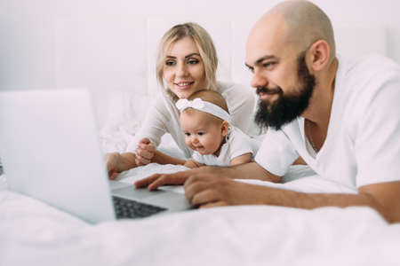 A beautiful Caucasian family mom, daughter and bearded dad lie on the bed in the bedroom in the morning and watch cartoons on a laptop. Parents and children.の写真素材