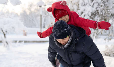 A man carries on his back a happy little girl a daughter in a red jacket, they play a game in the park in the snow in winter. Family concept.の写真素材