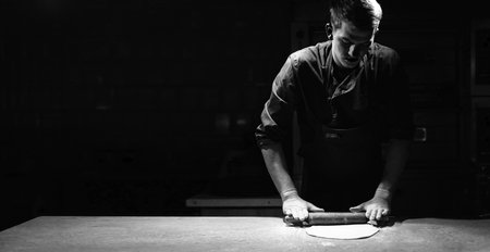 A male chef rolls out thin pizza dough on a table in a pizzeria. Banner and black background.の写真素材