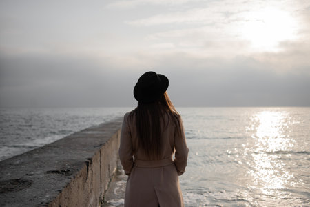 A young stylish girl in a light coat and hat stands on the seashore at the pier and looks into the distance. Business woman resting by the water after work.の写真素材