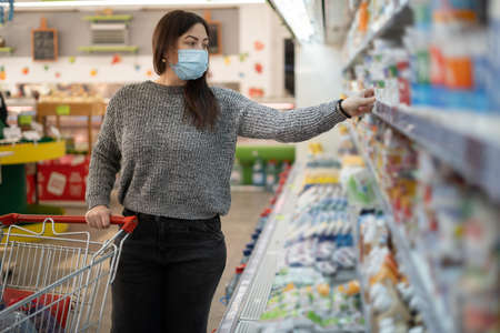 A girl in a protective mask stands with a cart near the refrigerators with dairy products in a grocery supermarket and takes milk from the shelf.の写真素材