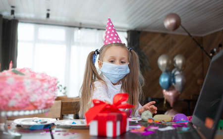 A schoolgirl in a medical mask and a festive cap stands near the table. The girl looks at the gift. Birthday celebration concept.の写真素材