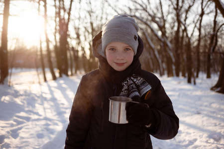 A child drinks hot chocolate or cocoa in a snowy winter park. Cute boy holding a cup of hot drink and smilingの写真素材