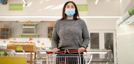 Young Caucasian woman in a medical mask stands in a supermarket. Shopping cart. Baner.