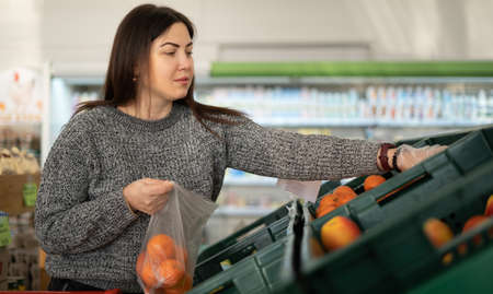 Young caucasian woman in a supermarket buys vegetables. Putting tangerines in a plastic bag. Citrus buying concept.の写真素材