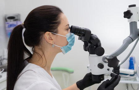 Dark-haired caucasian female dentist in a medical protective mask works with a modern microscope in...