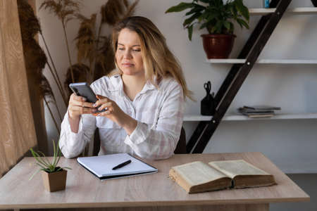 Caucasian girl reads SMS online in a smartphone sitting at a table at home with warm light from the window in the background. A student listens to a lecture on a smartphone and takes notes.の写真素材