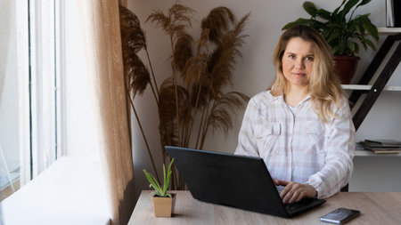 A pensive European woman with blond hair sits at a table and works on a laptop computer, looks to the future, thinks about solving problems in a home office, a serious girl is looking for inspiration.の写真素材