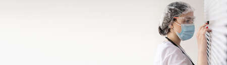 A young female doctor or nurse wearing a medical mask and glasses looks out the window, pulling the blinds apart with her fingers, and looks at something outside. Baner.の写真素材