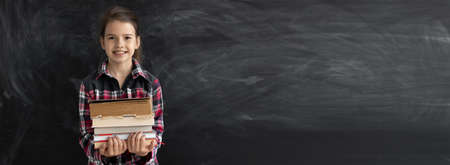 Caucasian girl student or schoolgirl in a shirt stands near a clean chalk board. holds old books in his hands. Back to school. copy space.の写真素材