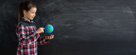 young caucasian girl schoolgirl or student stands near the chalk blackboard. Holds a globe. Banerの写真素材