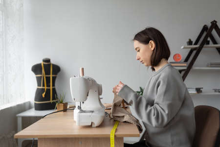 young caucasian woman fashion designer is sitting at home in her office at the table. Sewing machine. Measure the required piece of fabric with a flexible meter. Small business concept.の写真素材