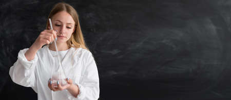 A scientist chemist in an office near a chalk board with a flask in his hands mixes liquids in a glass flask.Woman chemistry teacher shows the reaction to the students in the lesson.Black board.の写真素材