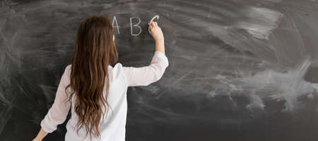 Rear view of a student or teacher with long brunette hair writing on a blank chalk board or blackboard with copyspace English alphabet. Back to school concept.の写真素材
