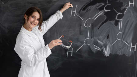 A young woman, a chemistry teacher or tutor, stands in a white coat near the blackboard and writes a formula. Back to school concept.の写真素材
