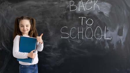 A young Caucasian girl schoolgirl stands against the background of a chalk board. The words are...