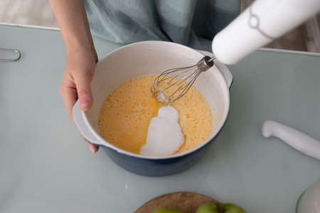 With an immersion blender with a whisk, the girl beats eggs with sugar in a ceramic saucepan. The process of making a dough for home baking. Cooking pies, cookies, pizza.の写真素材