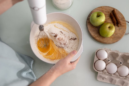 A girl chef in the kitchen kneads the dough with a blender with a whisk in a ceramic saucepan....