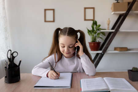 Caucasian girl schoolgirl sits at home at a desk. Talking on the phone and making notes in a...