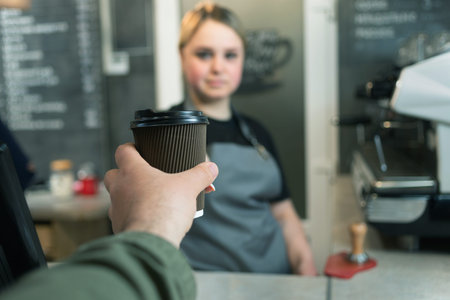 Close-up of a shopper customers hand with a paper glass of hot espresso coffee in a cafe and a...