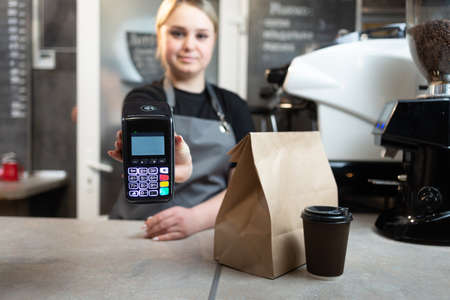 Contactless payment of an order through a payment terminal with nfs to a barista girl in a cafe for coffee and food in paper packaging.The girl holds out the device to pay for the order.の写真素材