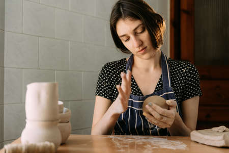 a young charismatic woman with long black hair sits at home and sculpts a clay pot. A large piece of clay is kneading in his hands.の写真素材